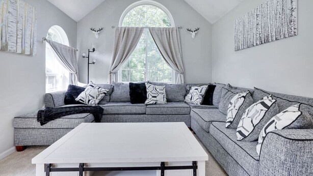 Un salon moderne avec un canapé d’angle gris, une table basse blanche et une grande fenêtre avec des rideaux.