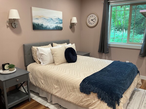 A bedroom with a bed, nightstands, a clock, and a window with a view of greenery.