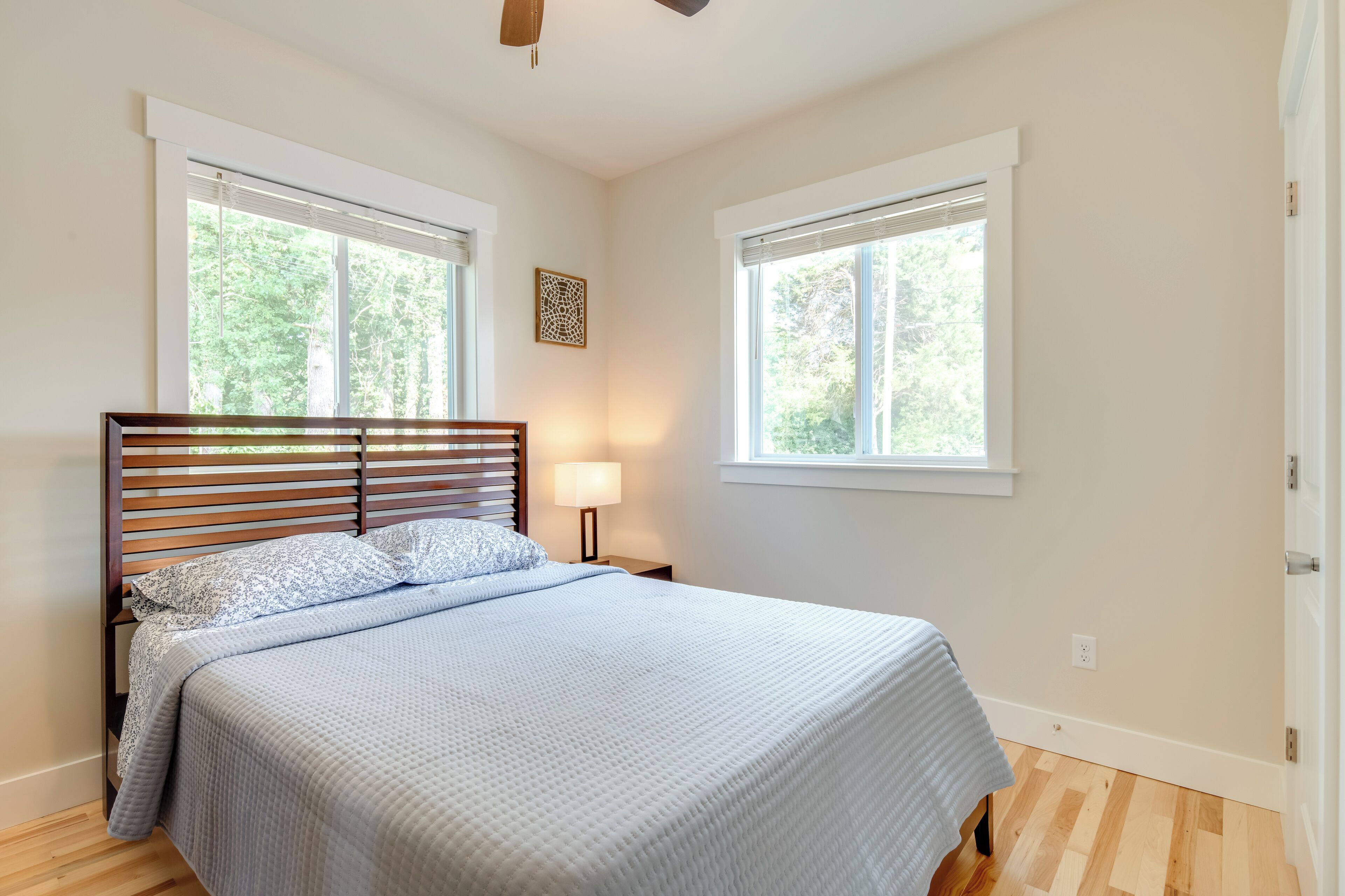 A bedroom with a wooden bed, two windows with blinds, a bedside lamp, and a small decorative wall piece.