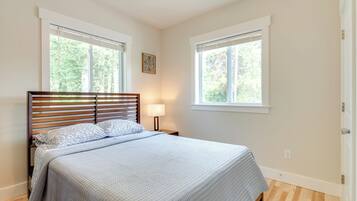 A bedroom with a wooden bed, two windows with blinds, a bedside lamp, and a small decorative wall piece.