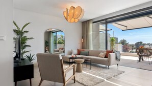 A modern living room with a sofa, armchair, and coffee table, a large window with a view, and a unique ceiling light fixture. - Casa Hermano - Private Pool Penthouse (Marbella)