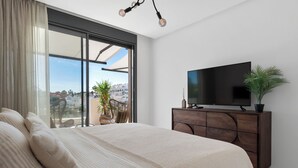 A bedroom with a large bed, a TV on a wooden cabinet, and a view of the city through large windows. - Casa Hermano - Private Pool Penthouse (Marbella)