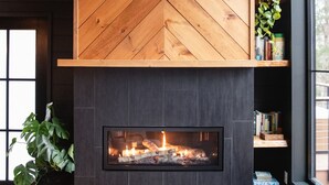 A modern interior with a herringbone-patterned wooden wall above a fireplace, dark tiled lower section, and a window with a view.