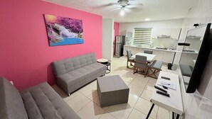 A living room with a grey sofa, a dining table with chairs, a kitchen area with a fridge, and a ceiling fan.
