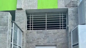 A modern building entrance with a white door, a small window with metal bars, and a green wall above.