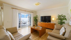 Een moderne woonkamer met een zithoek, een salontafel, een televisie op een houten kast, en een schuifdeur naar een buitenruimte.