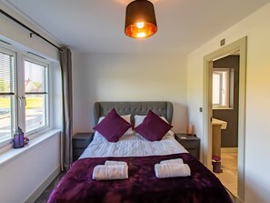 Une chambre d’hôtel avec un lit, deux oreillers, une fenêtre, une table de chevet et une salle de bain visible à travers une porte ouverte.