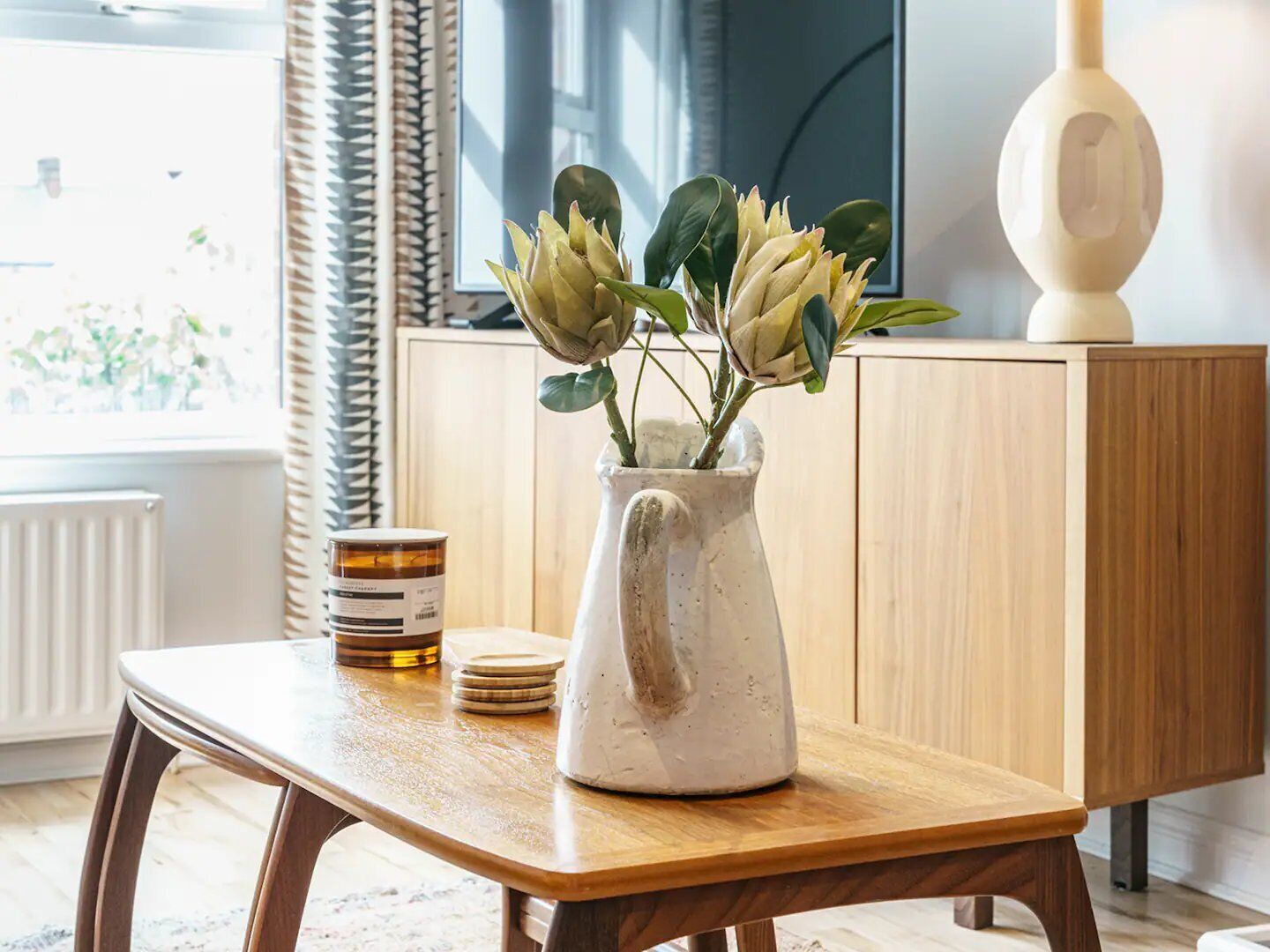 A wooden table with a white ceramic pitcher containing green plants, a stack of coasters, and a jar of honey.
