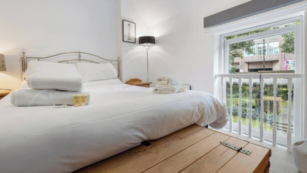 A neatly made bed with white linens, a bedside lamp, and a window with a view of a building and greenery outside. - 4, 35 Fossgate · Cosy Central 2-Bed Home Right in the Heart of York - Pass the Keys (York)