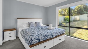 A bedroom with a large bed, white nightstands, a wooden headboard, and a view of a backyard through a sliding glass door. - Tura Beach Getaway on Telopea (Tura Beach)