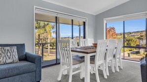 A balcony with a dining table and chairs, a sofa, and large windows offering a view of trees and a distant landscape. - Tura Beach Getaway on Telopea (Tura Beach)