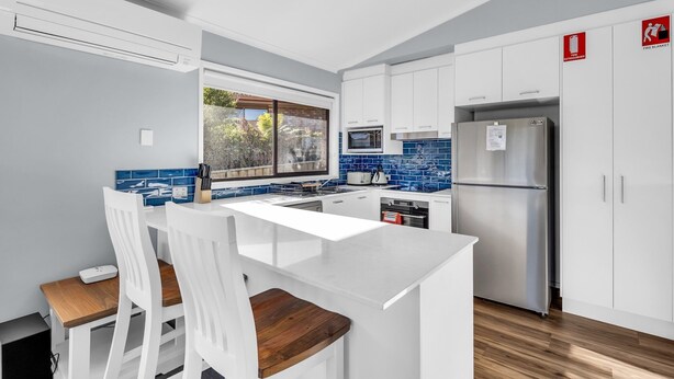 A modern kitchen with white cabinets, a stainless steel refrigerator, a microwave, and a large window overlooking a garden. - Tura Beach Getaway on Telopea (Tura Beach)
