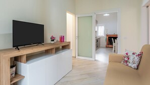 A living room with a TV on a wooden stand, a sofa with a floral pillow, and a kitchen in the background.