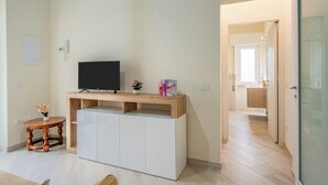 A modern living room with a flat-screen TV on a wooden console, a small round table with a plant, and a hallway leading to a kitchen.