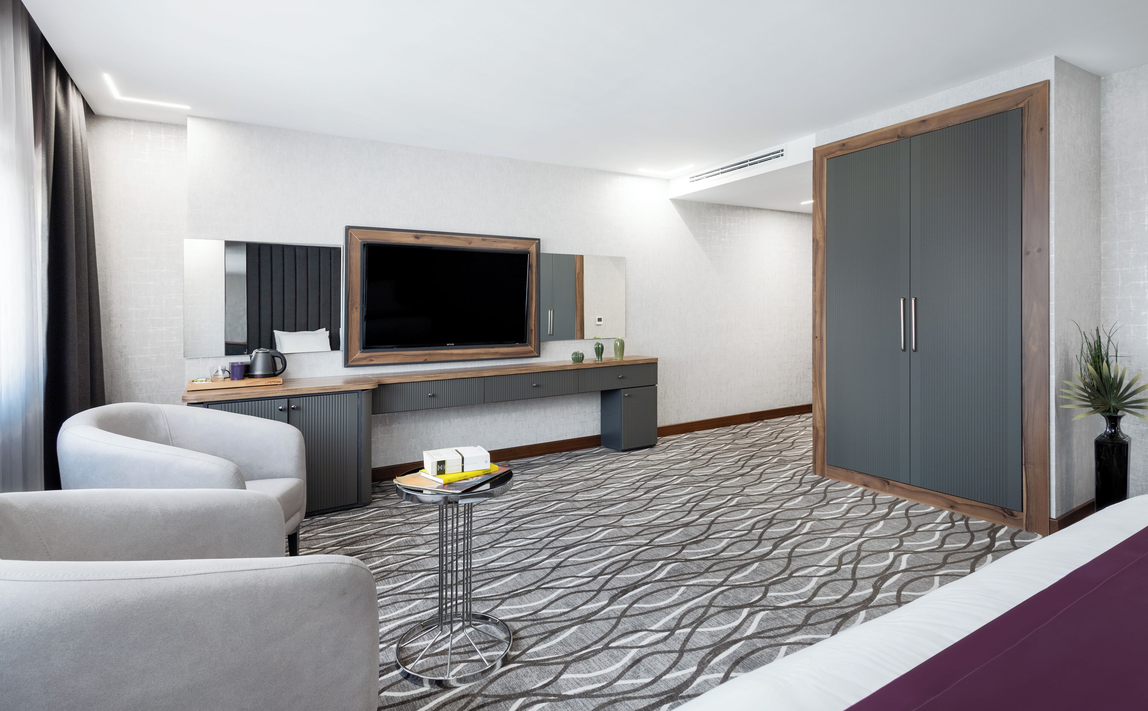 A modern hotel room with a large bed, a desk with a flat-screen TV, a seating area with two chairs, and a wardrobe.
