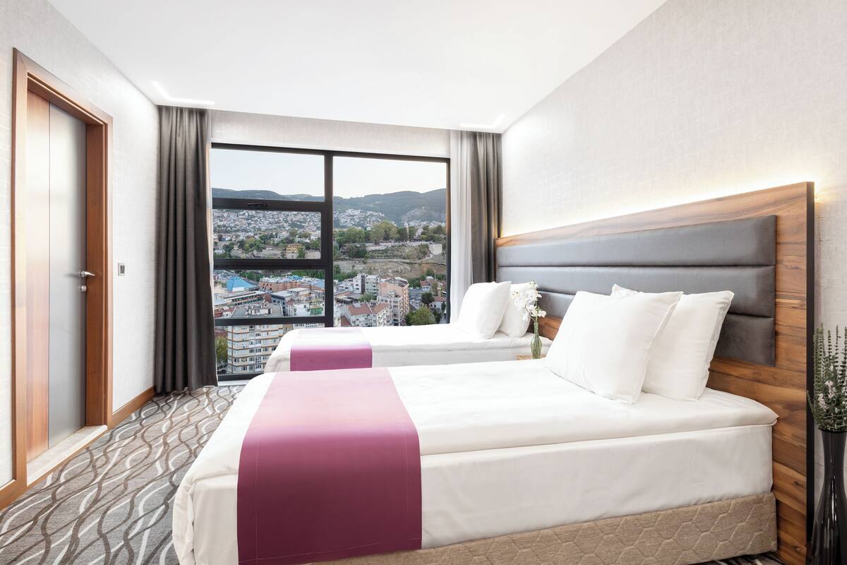 A hotel room with two beds, a large window with a city view, and a wooden headboard.