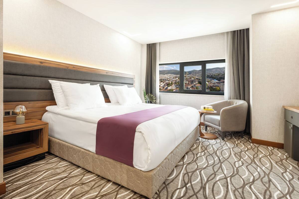 A hotel room with a large bed, a small desk, a chair, and a window with a city view.