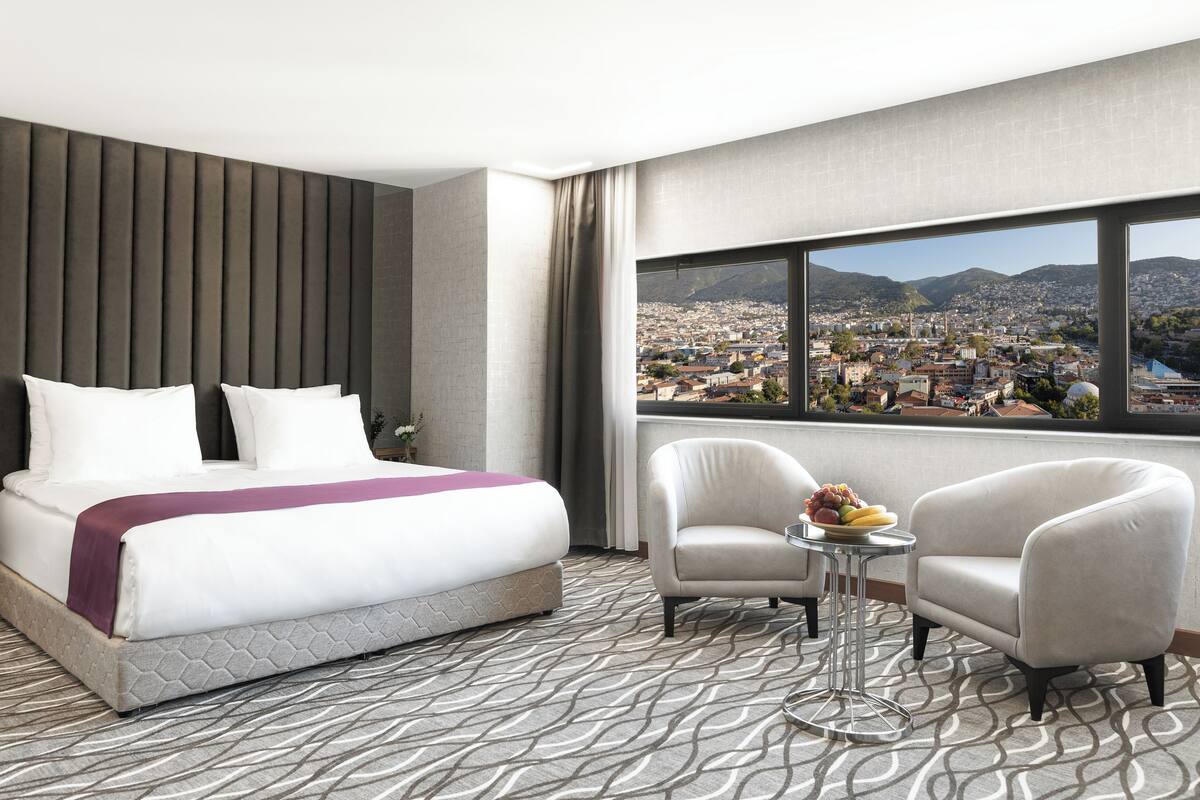 A hotel room with a large bed, two armchairs, a small table with fruit, and a city view through large windows.