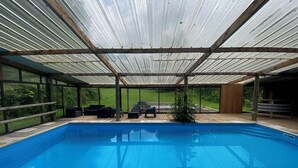 An indoor swimming pool with a wooden deck and seating area, surrounded by glass walls and a metal roof.