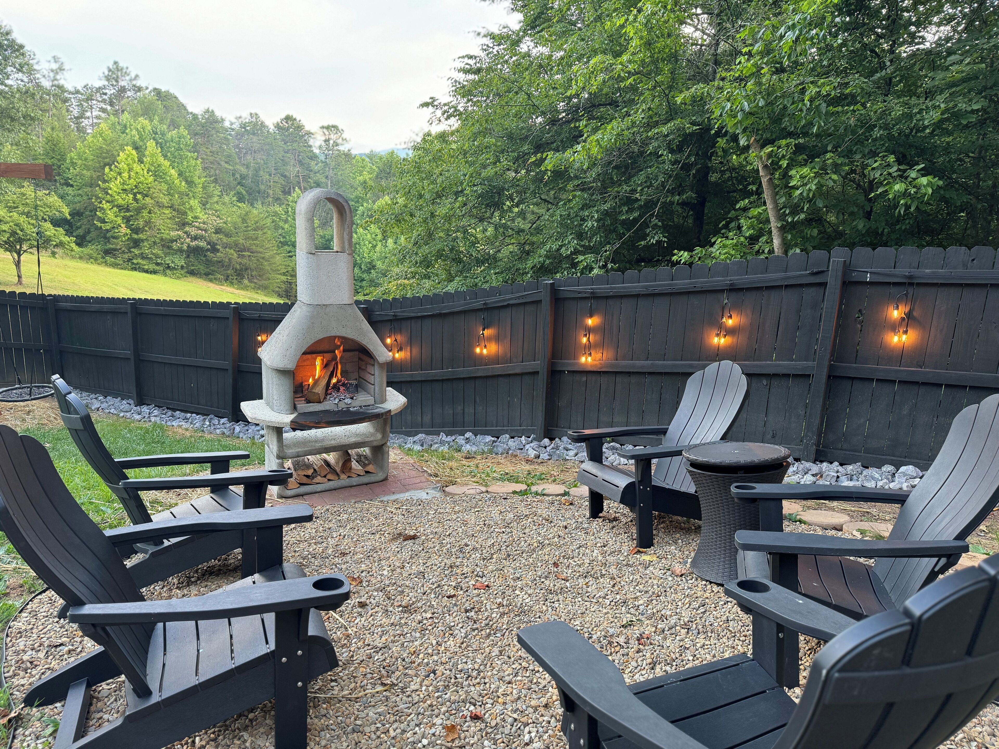 A backyard with a stone fire pit, Adirondack chairs, and a wooden fence.