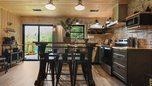 A modern kitchen with wooden cabinets, a central island, bar stools, and a view of greenery outside. - Smokies Pond Escape/Secluded Mtn cabin w/pond/Fish’n/Hot tub/2 fenced yards/Pets (Pigeon Forge)