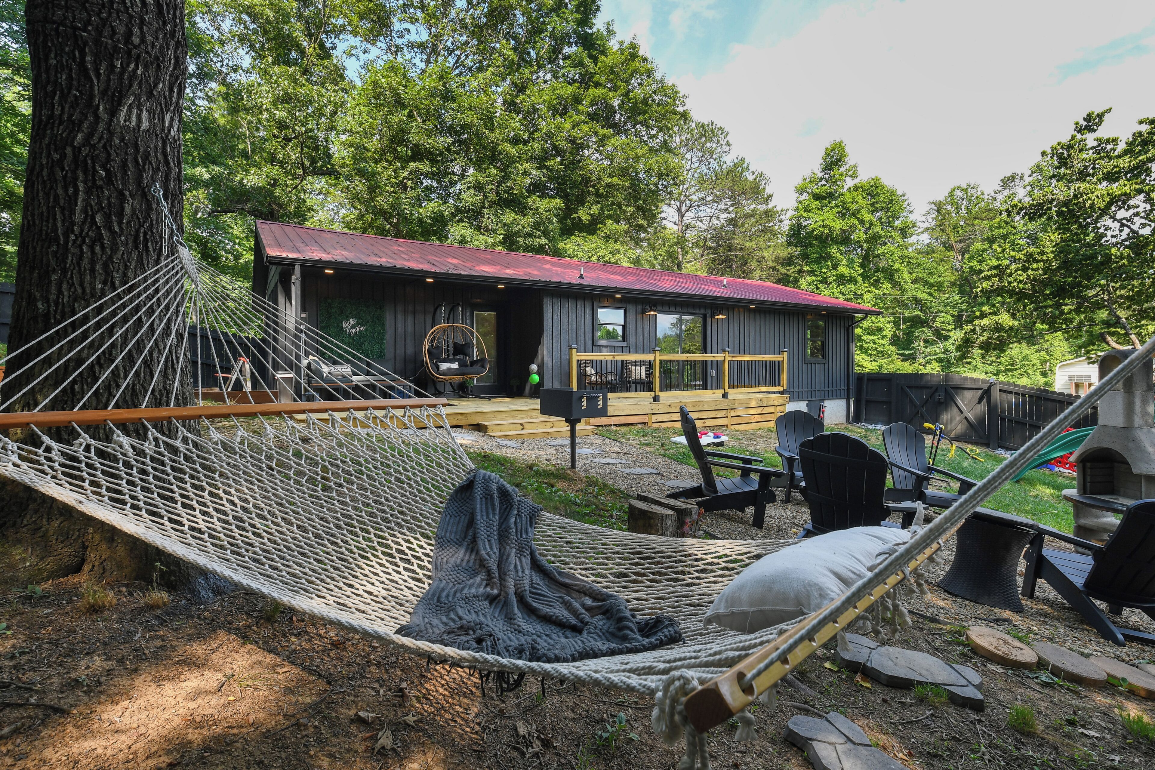 A hammock tied to a tree, a wooden deck, outdoor chairs, and a small building with a red roof.