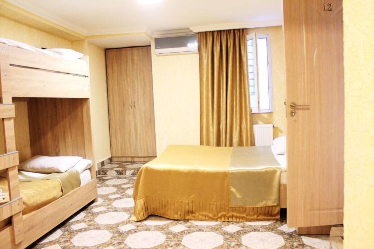 A room with a bunk bed, a single bed with a gold cover, a wooden wardrobe, a door with the number 12, and a patterned floor.