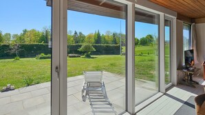 Una habitación con grandes puertas corredizas de vidrio que dan a un espacio exterior con césped, una banca y un televisor sobre un pedestal de madera.