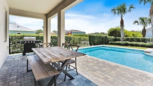 Un área de piscina al aire libre con mesa y bancos de madera, rodeada por un seto y palmeras.