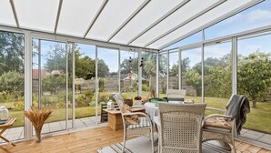 Een oranjerie met rieten meubilair, een houten tafel en uitzicht op een tuin.