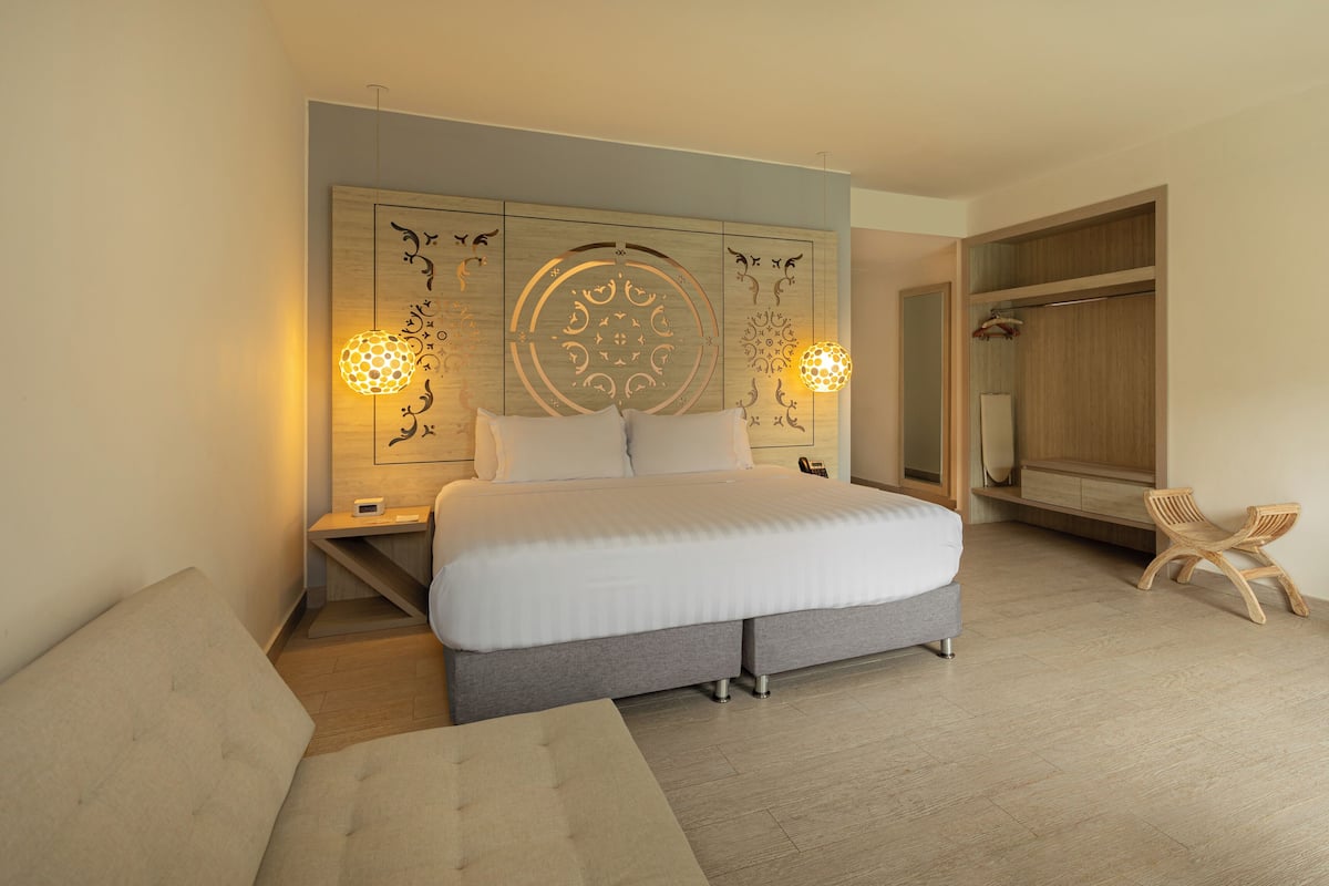 A modern hotel room with a large bed, a bedside table, a lamp, a wardrobe, and a small seating area.