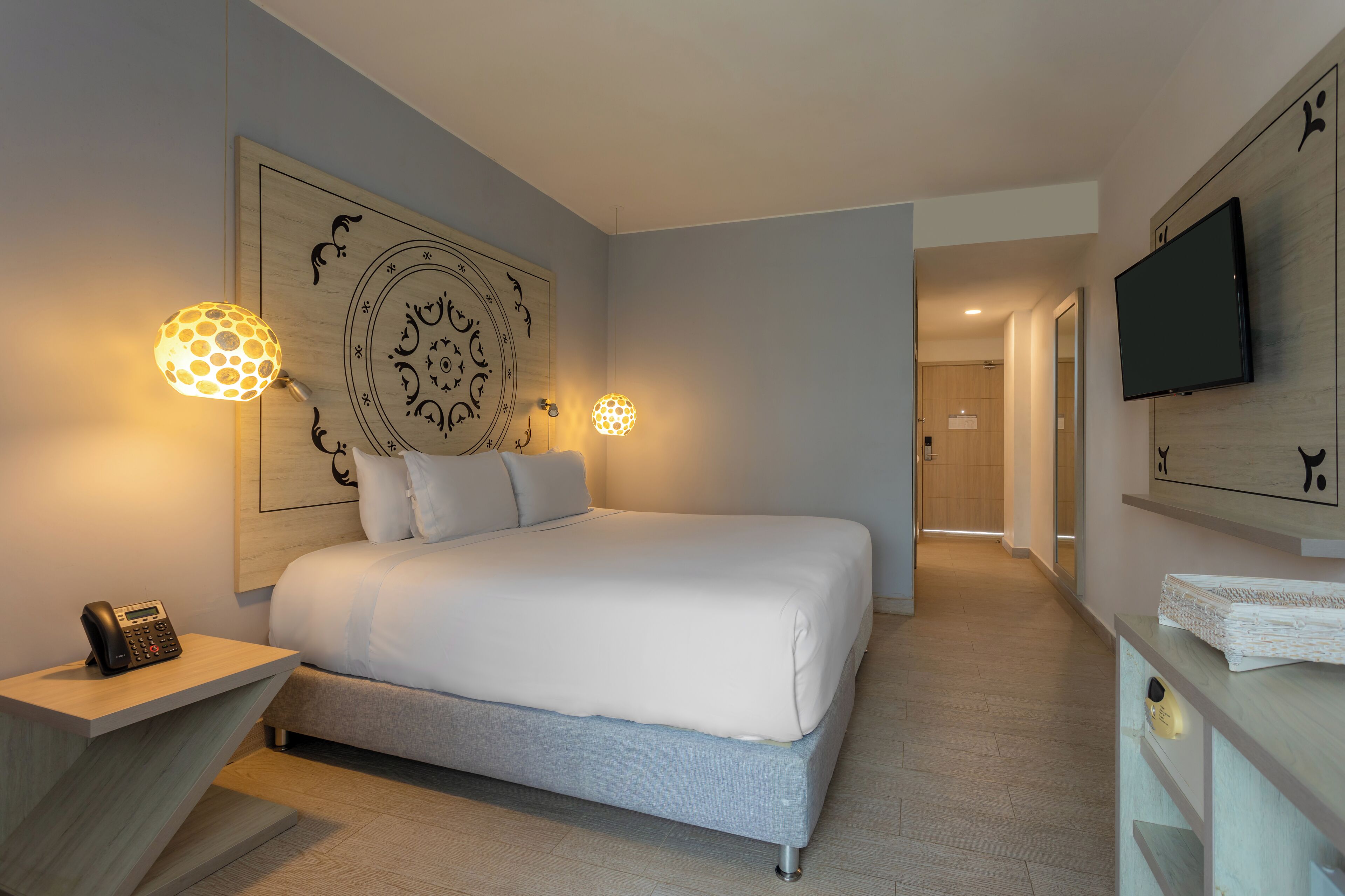 Une chambre d’hôtel moderne avec un grand lit, deux lampes fixées au mur, un téléviseur à écran plat et une tête de lit en bois.