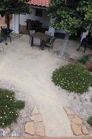 En udendørs patio med et bord og stole, omgivet af en grussti og en lille have med hvide blomster.