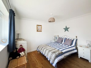 Um quarto com uma cama, mesa de cabeceira, cadeira de vime, janela com cortinas, um adorno de estrela na parede e uma moldura com imagem.