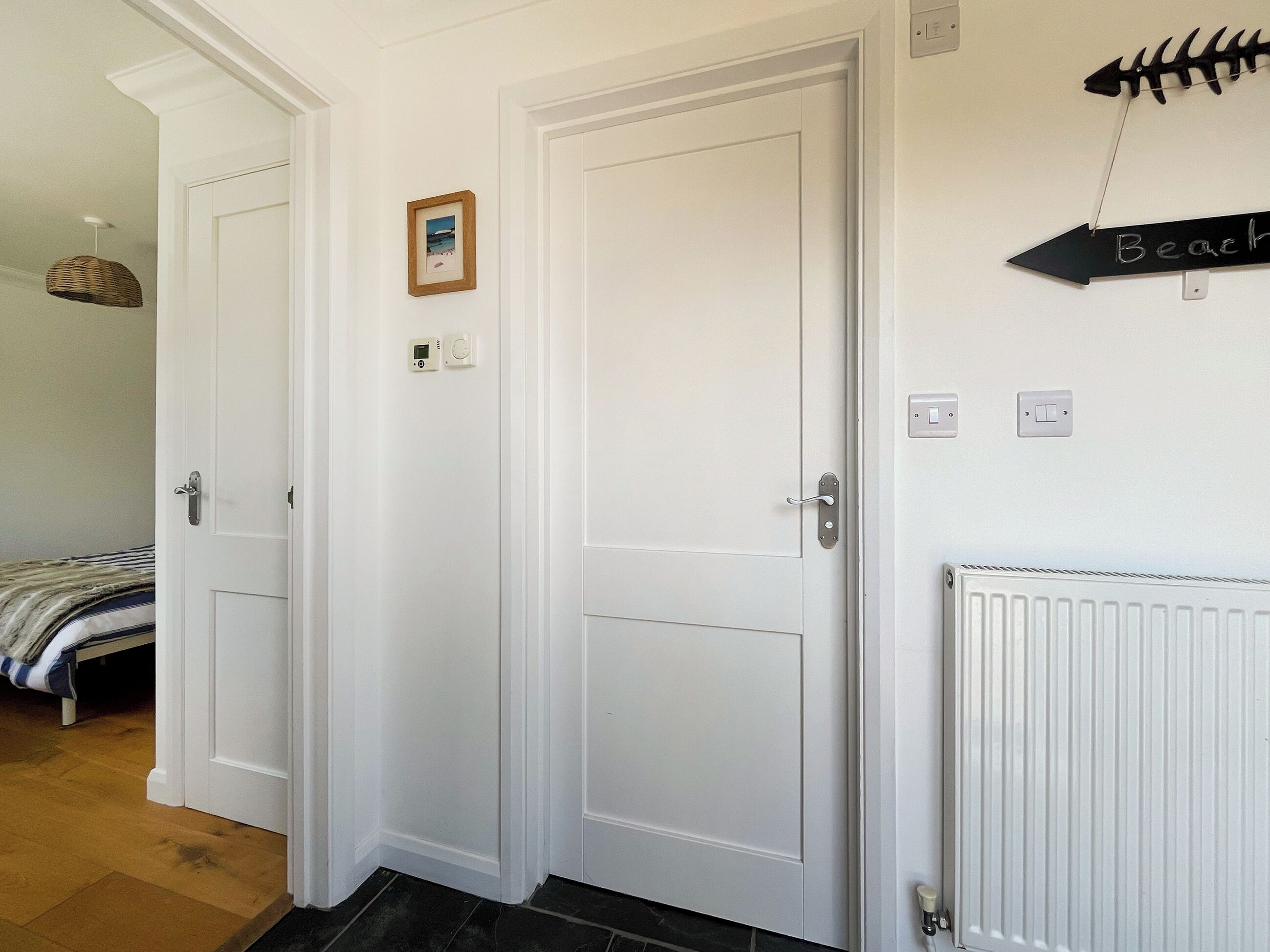 Una habitación con dos puertas blancas, un pequeño cuadro en la pared, un termostato y un cartel que indica el camino a la playa.