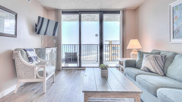 A balcony with a view of the beach, a sofa, a chair, a TV, and a lamp. - Ocean Trillium 803 - a 1 bedroom condo with beautiful ocean views (New Smyrna Beach)