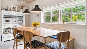 Un comedor con mesa de madera, sillas a rayas y un jarrón con girasoles. Dos lámparas colgantes iluminan la mesa, y una ventana ofrece vistas a la vegetación exterior.
