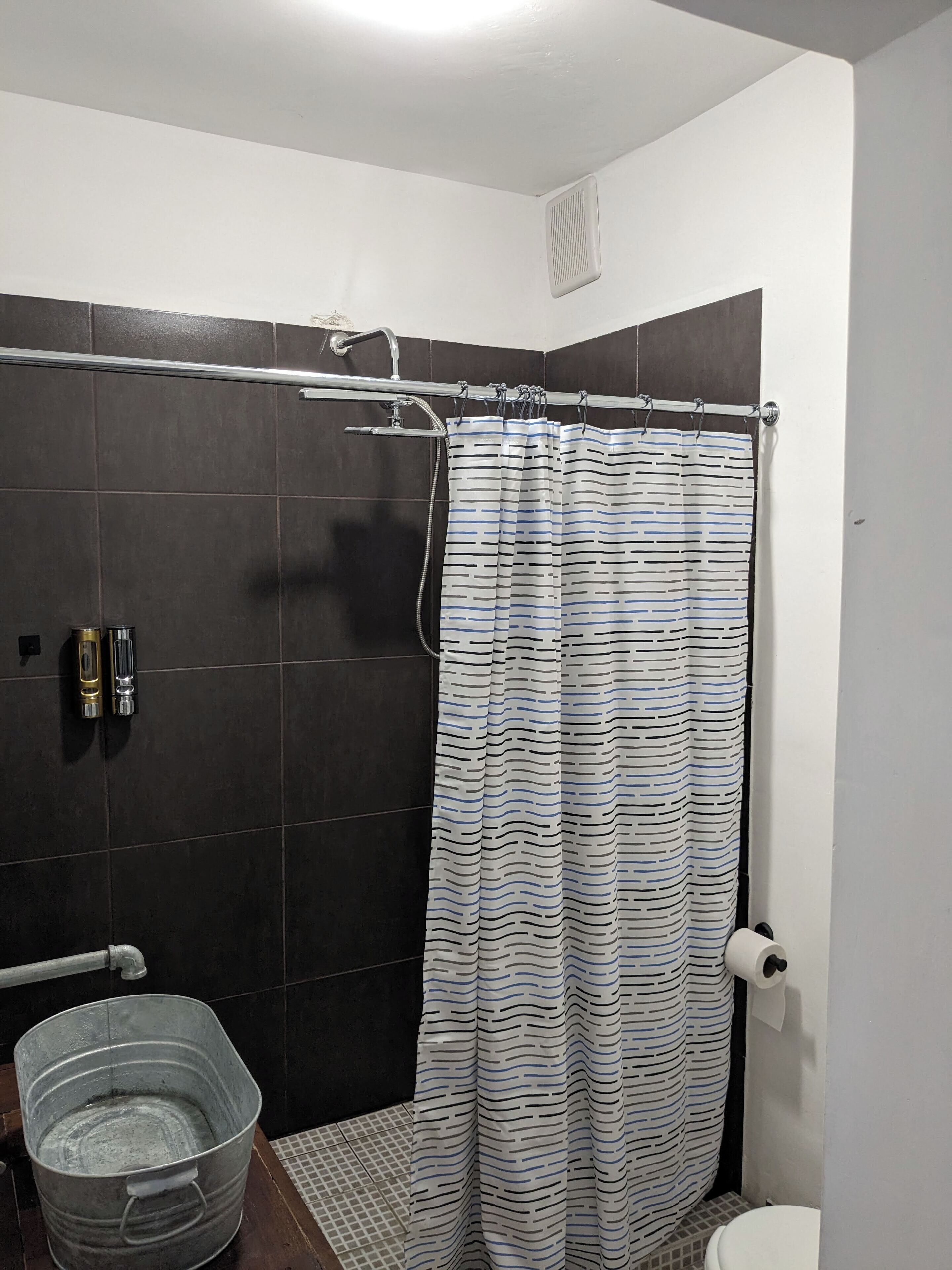 A bathroom with a shower, dark tiled walls, a striped shower curtain, a metal bucket, and a toilet.
