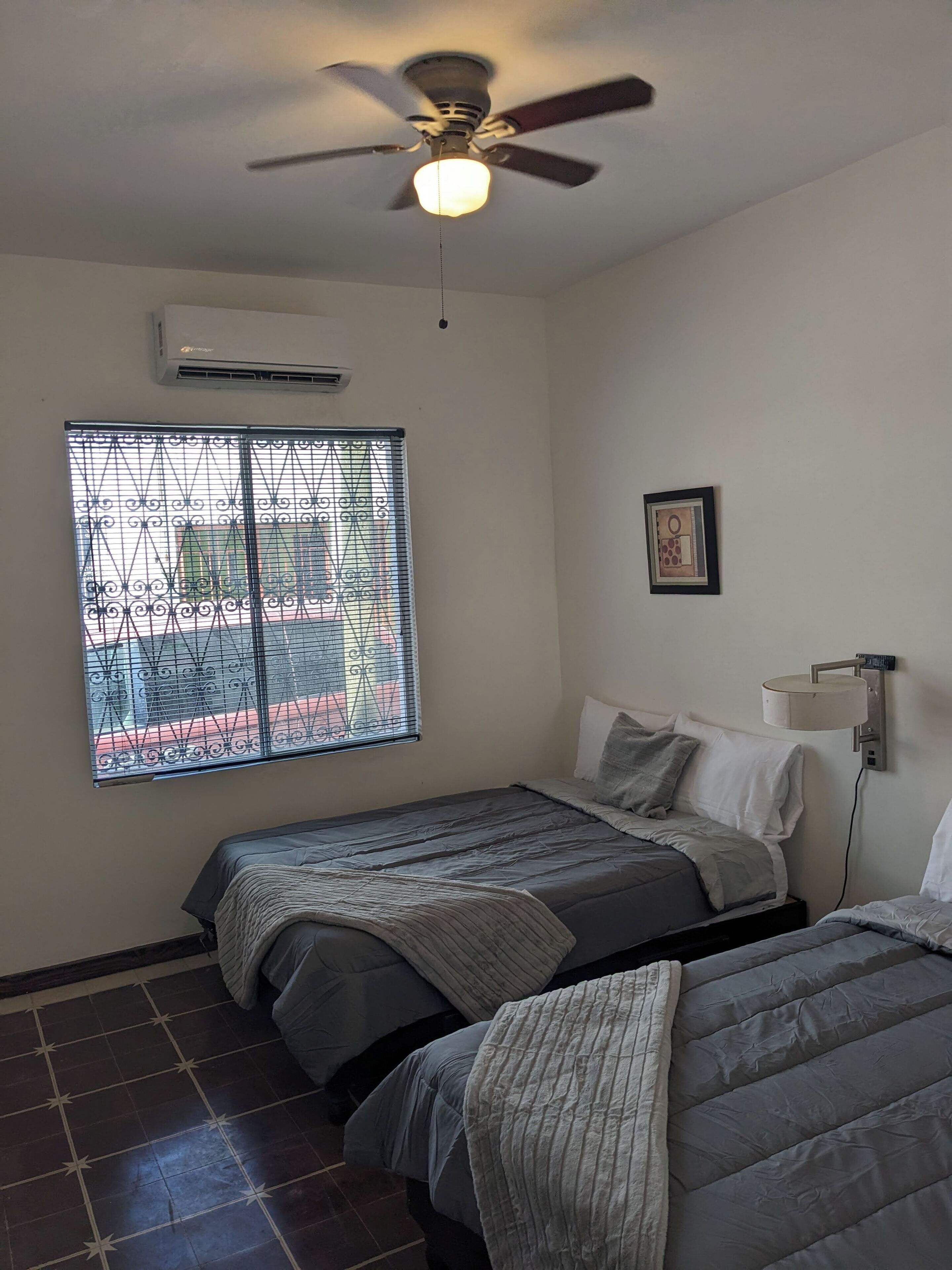 A bedroom with two beds, a ceiling fan, an air conditioner, a window with metal bars, and a wall-mounted lamp.