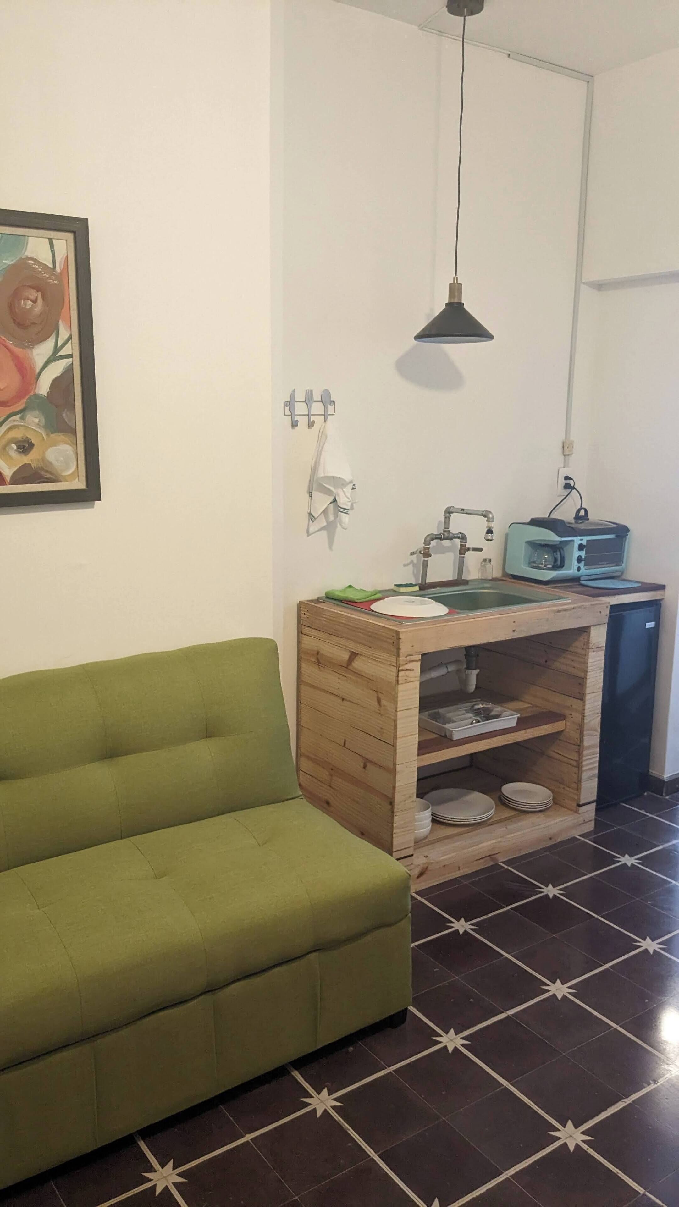 Un espacio habitable compacto con un sofá verde, una kitchenette con tostadora y una lámpara colgante.