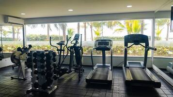 Un gimnasio con cintas de correr, pesas y una bicicleta estática, con grandes ventanales que dan a palmeras.