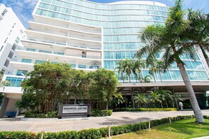 Un hotel moderno con fachada de vidrio curva, varios balcones y exuberante jardinería, incluyendo palmeras y setos bien cuidados.