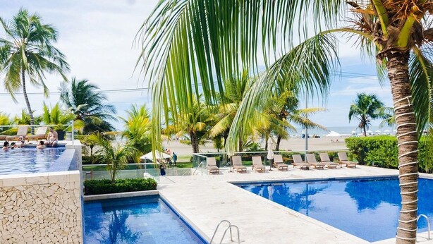 A pool area with two swimming pools, lounge chairs, palm trees, and a view of the beach. - U-GO Edificio Morros Ultra (Cartagena)