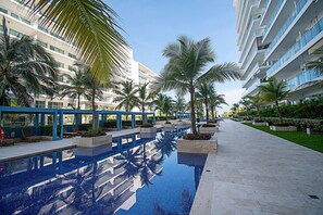 Zona de piscina con palmeras, cielo despejado y edificios modernos al fondo.