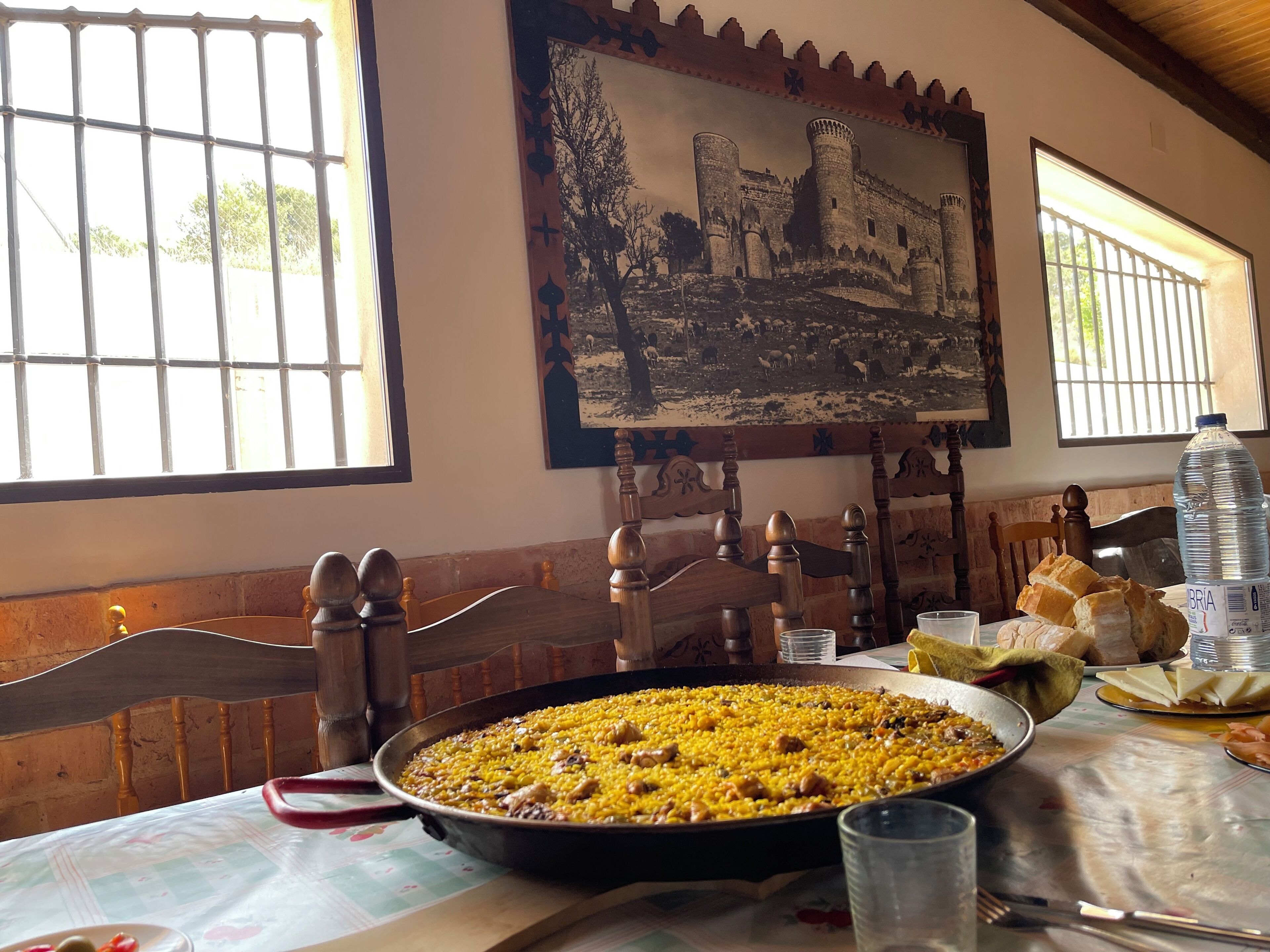Taula de menjador amb una paella gran, cadires, un quadre i una finestra amb reixes.