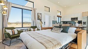 Ett modernt hotellrum med en stor säng, en sittgrupp med en fåtölj och ett bord, ett litet kök och utsikt över ökenlandskapet.