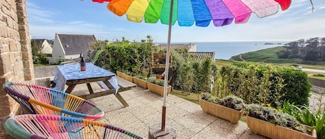 Een terras met tafel en stoelen, een kleurrijke parasol en uitzicht op zee.