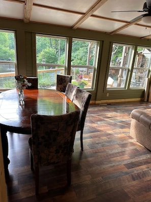 A dining area with a large wooden table and patterned chairs, large windows with a view of greenery, and a ceiling fan.