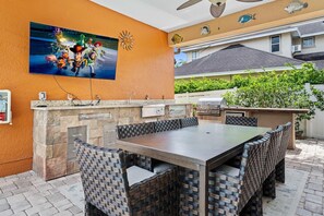 An outdoor kitchen with a stone island, dining table, and chairs, surrounded by a patio area with a view of a residential neighborhood.