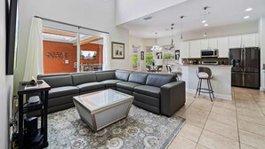 A modern living room with a sectional sofa, a glass coffee table, and a kitchen area with stainless steel appliances. - Theater Room | Games Area | Kids Themed Bedrooms | Private Pool (Kissimmee)